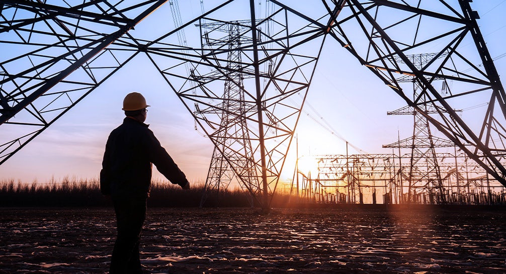 A man in a hard hat is standing in front of a high voltage tower.