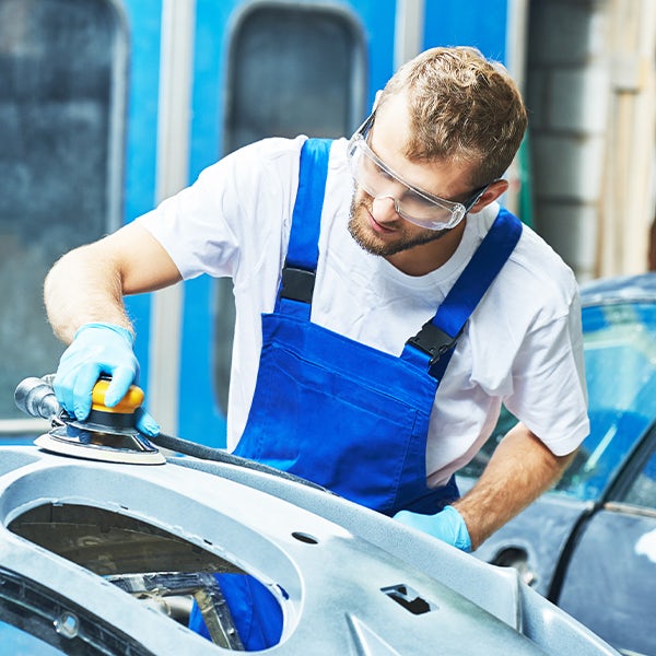 A man wearing blue overalls is sanding a car.