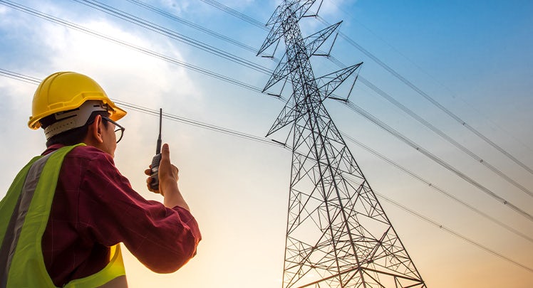 A man in a hard hat is holding a radio in front of high voltage pylons.