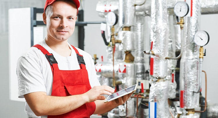 A plumber holding a tablet in front of pipes.