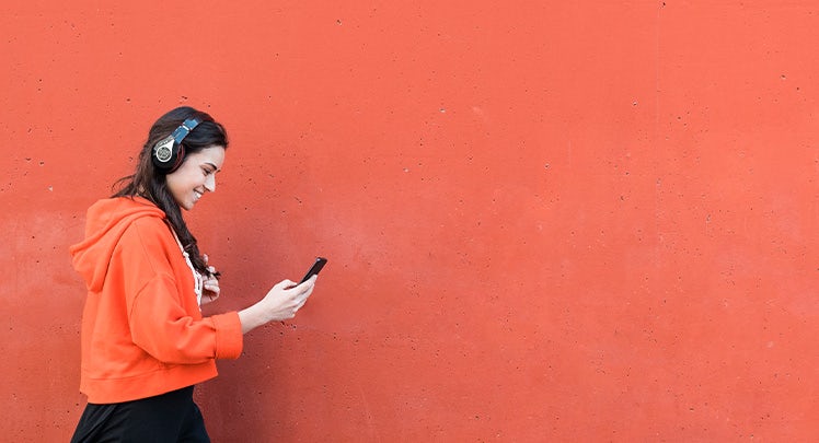 A woman wearing an orange hoodie and headphones is standing against an orange wall.