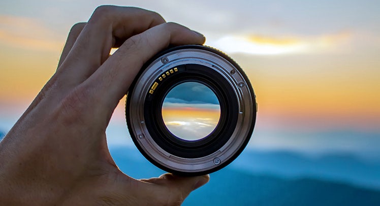 A person holding up a camera lens with a sunset in the background.