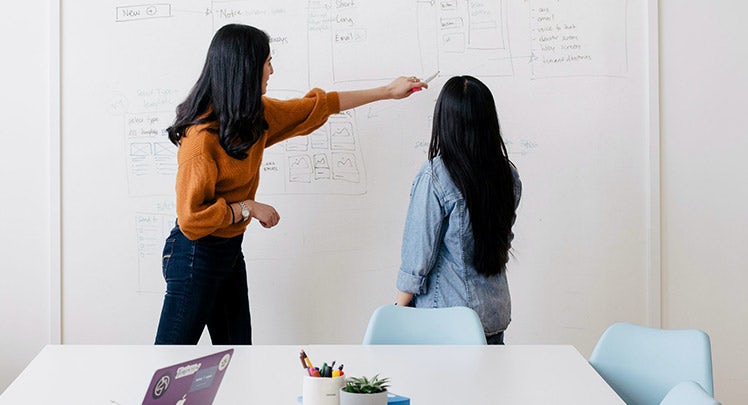 Two women pointing at a whiteboard in an office.