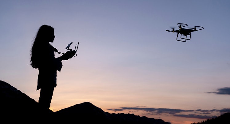 A silhouette of a woman flying a drone at sunset.