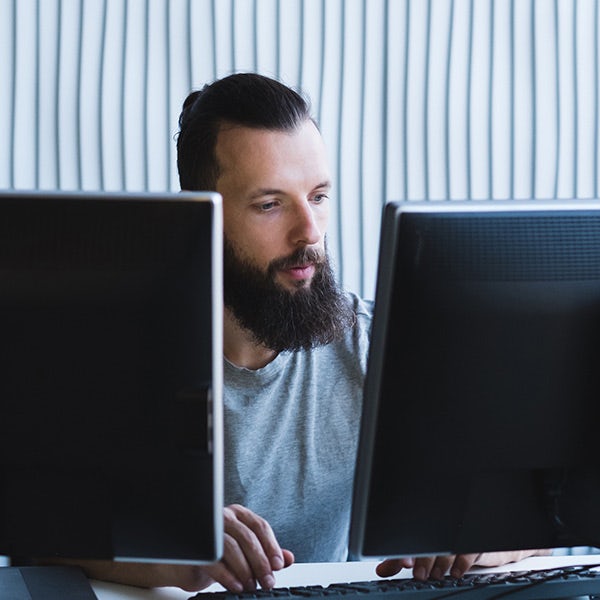 A bearded man sitting in front of two computer monitors.