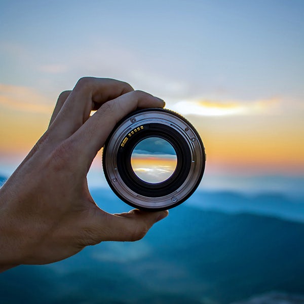 A person holding up a camera lens at sunset.